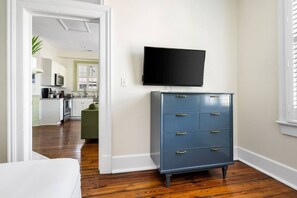 A room with a wooden floor, a blue dresser with drawers, a flat-screen TV mounted on the wall, and a doorway leading to a kitchen area. - 2BR Spring Street Retreat - B (Charleston)