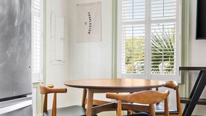 Et køkken med et rustfrit stål køleskab, et træbord med stole, et vindue med persienner og et TV ophængt på væggen.
