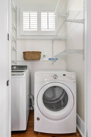 A laundry room with a front-loading washing machine and a dryer, shelves above for storage, and a window with blinds. - 2BR Spring Street Retreat - B (Charleston)