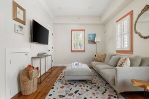 A living room with a grey sofa, a coffee table, a TV, and a window with shutters. - 1 BR Cottage with Parking - Charleston Charm (Charleston)