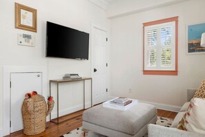 A living room with a flat-screen TV mounted on the wall, a small table with books, a wicker basket, and a grey sofa. - 1 BR Cottage with Parking - Charleston Charm (Charleston)