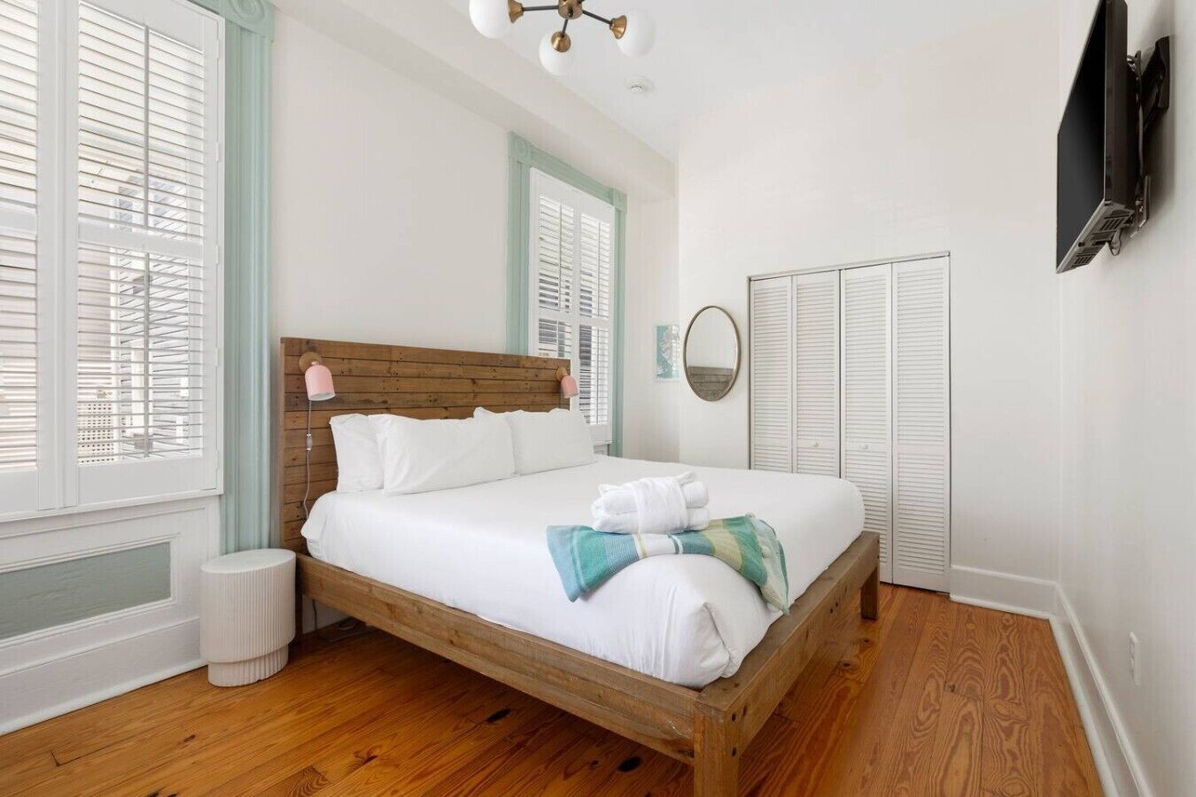 A neatly made bed with white linens and a blue and white throw blanket, wooden headboard, white walls, and a wooden floor.