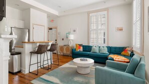A modern living room with a teal sofa, a coffee table, and a kitchen area with a refrigerator and bar stools. - 2BR Spring Street Escape - D (Charleston)