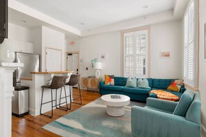 Ein modernes Wohnzimmer mit einem türkisfarbenen Sofa, einem Couchtisch und einer Küchenzeile inklusive Kühlschrank und Barhocker.