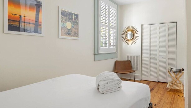 A bedroom with a bed, a chair, a wardrobe, and two framed artworks on the wall. - 2BR Spring Street Escape - D (Charleston)
