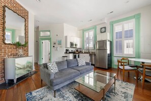 A modern living room with a grey sofa, a glass coffee table, and a dining area with wooden chairs. - 8BR - Beautiful CHS Home | Great for Groups (Charleston)