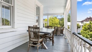 A covered porch with a dining table and chairs, white railings, and a view of greenery and buildings in the background. - 2BR Historic Charleston Getaway - C (Charleston)