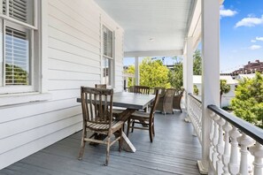 Eine überdachte Veranda mit Esstisch und Stühlen, weißen Geländern und Blick auf Grünflächen und Gebäude im Hintergrund.