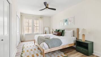 Un dormitorio con cama, mesitas de noche, ventilador de techo y ventana con cortinas.
