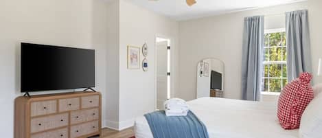 A bedroom with a bed, a television on a wooden dresser, a ceiling fan, and a window with curtains.