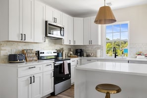 A modern kitchen with white cabinetry, a large island, a microwave, and a hanging pendant light. - 4BR New - Close to Folly Beach! (Charleston)