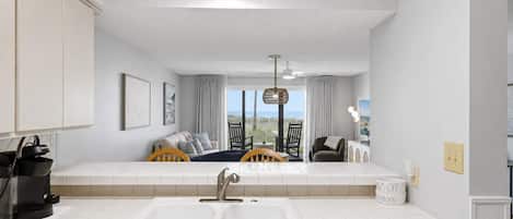 A modern kitchen with a large window, a dining area with a view, and a living space with a sofa and a TV.