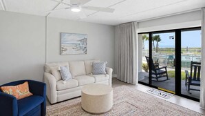 Ein Wohnzimmer mit einem weißen Sofa, einem blauen Sessel, einem runden Hocker, einem Deckenventilator und einer großen Glasschiebetür, die auf einen Balkon mit Gartenmöbeln führt.