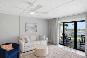 Sala de estar con sofá blanco, sillón azul, otoman redonda, ventilador de techo y un amplio ventanal corredera que da a un balcón con muebles de exterior.