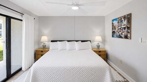 Ein Hotelzimmer mit einem Bett, zwei Nachttischen mit Lampen, einem Deckenventilator, einem großen Fenster mit Vorhängen und einem Wandbild.