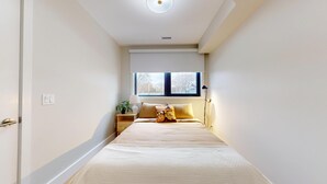 A narrow hotel room with a bed, a nightstand, a window, and a ceiling light.