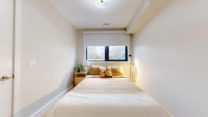 A narrow hotel room with a bed, a nightstand, a window, and a ceiling light.