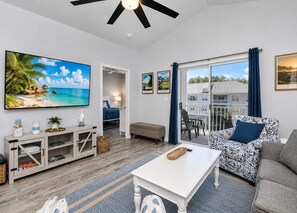 Ein modernes Wohnzimmer mit einem Flachbildfernseher, einer Couch, einem Couchtisch und einem Balkon mit Blick auf ein Haus.