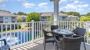 A covered balcony with wicker furniture and a glass table overlooking a swimming pool and residential buildings. - Golf, Pool & Coastal Comfort Willow Bend 234 (North Myrtle Beach)