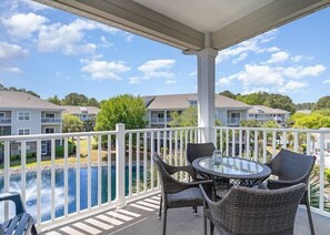Ein überdachter Balkon mit Korbmöbeln und einem Glastisch mit Blick auf einen Pool und Wohnhäuser.