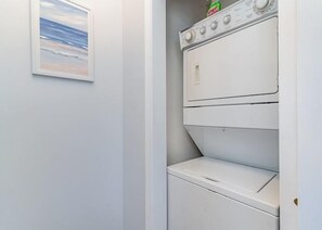 Ein Waschraum mit übereinander gestapelten Waschmaschinen und Trocknern, einem gerahmten Bild an der Wand und einer Tür zu einem anderen Raum.