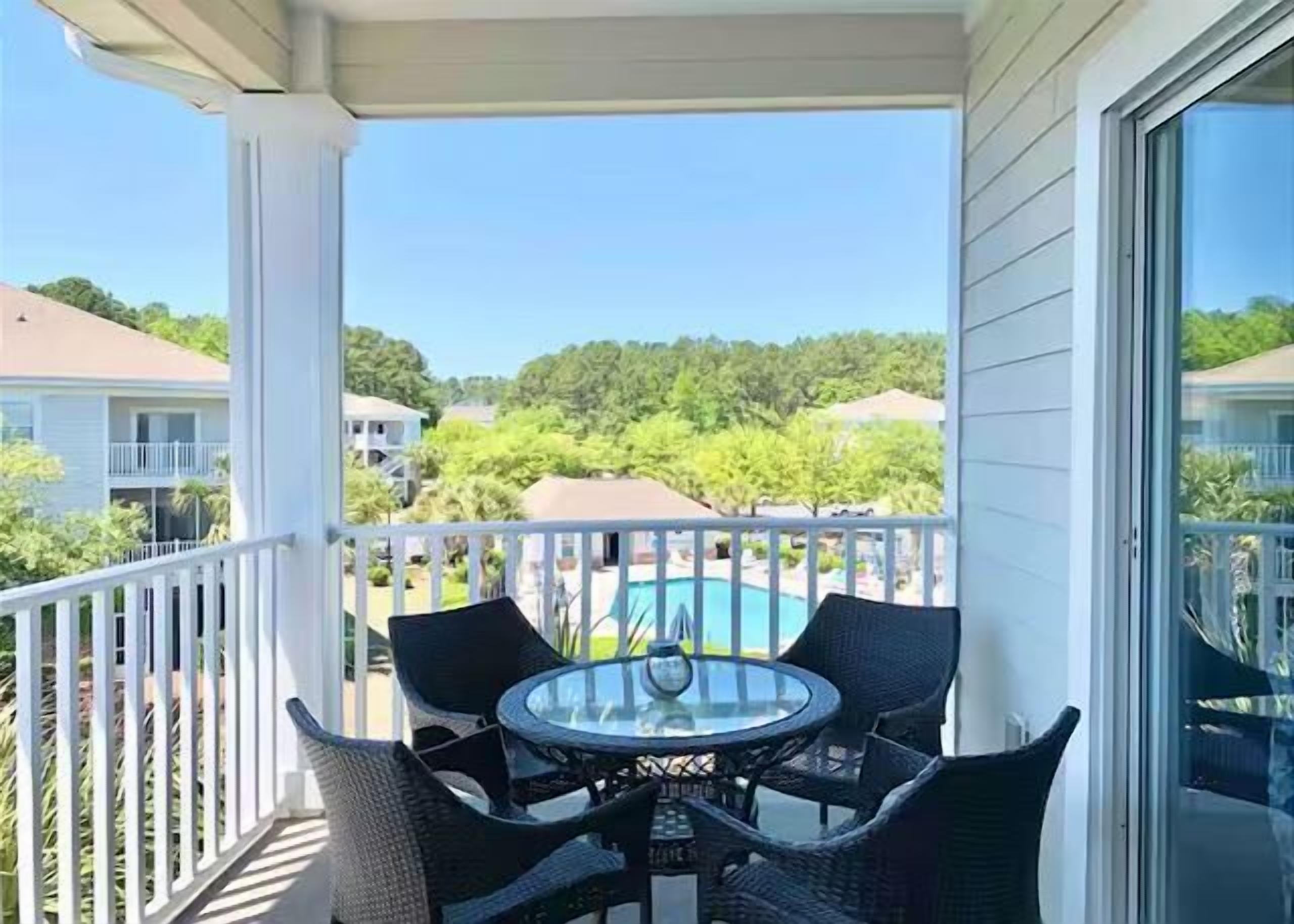 A balcony with a table and chairs, overlooking a swimming pool and greenery.
