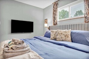 Habitación de hotel con cama, televisor de pantalla plana montado en la pared, ventana con cortinas y mesita de noche con toallas.