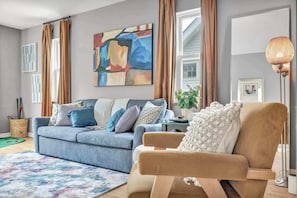 A living room with a blue sofa, a beige armchair, a colorful rug, and a large abstract painting on the wall.