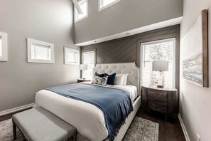 A bedroom with a large bed, a nightstand, a bench, and a window with blinds. - East Nashville Social House | Sleeps 10 + Deck (Nashville)