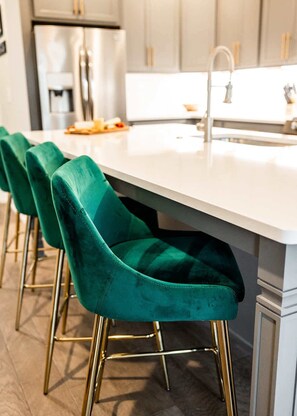 A kitchen with a white countertop, a stainless steel refrigerator, and a sink. There are green upholstered bar stools with gold legs. - East Nashville Social House | Sleeps 10 + Deck (Nashville)