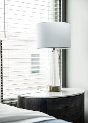 A modern lamp with a white shade on a bedside table next to a window with blinds. - East Nashville Social House | Sleeps 10 + Deck (Nashville)