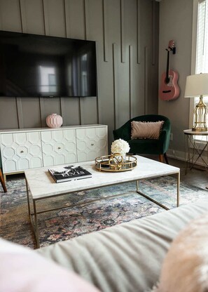A living room with a flat-screen TV mounted on the wall, a green armchair, a coffee table with decorative items, and a patterned rug. - East Nashville Social House | Sleeps 10 + Deck (Nashville)