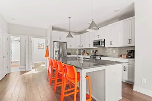 A modern kitchen with white cabinets, a stainless steel refrigerator, a microwave, and a large island with orange bar stools. - Nashville Pop Art 4BR, Near Downtown and Sleeps 10 (Nashville)
