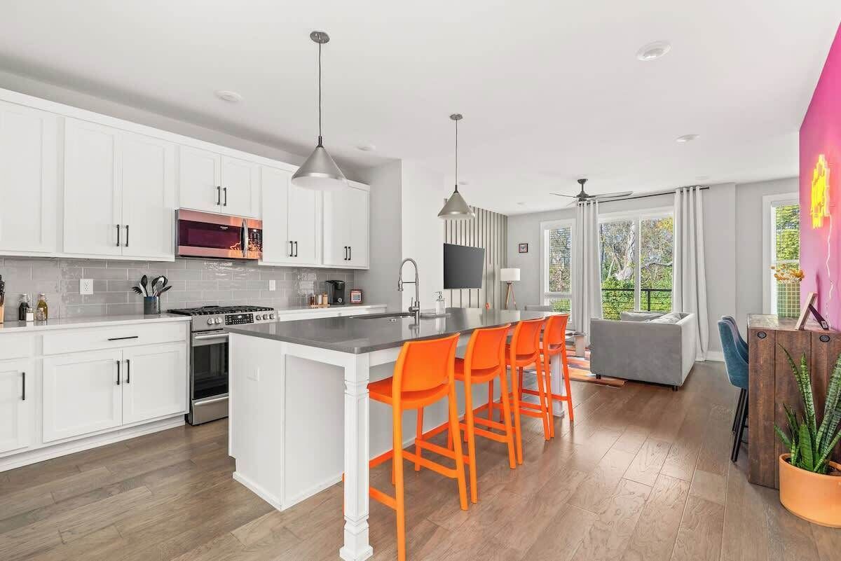 A modern kitchen with white cabinets, a central island with orange chairs, a microwave, stove, and a large window offering a view of greenery.