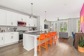 A modern kitchen with white cabinets, a central island with orange chairs, a microwave, stove, and a large window offering a view of greenery. - Nashville Pop Art 4BR, Near Downtown and Sleeps 10 (Nashville)