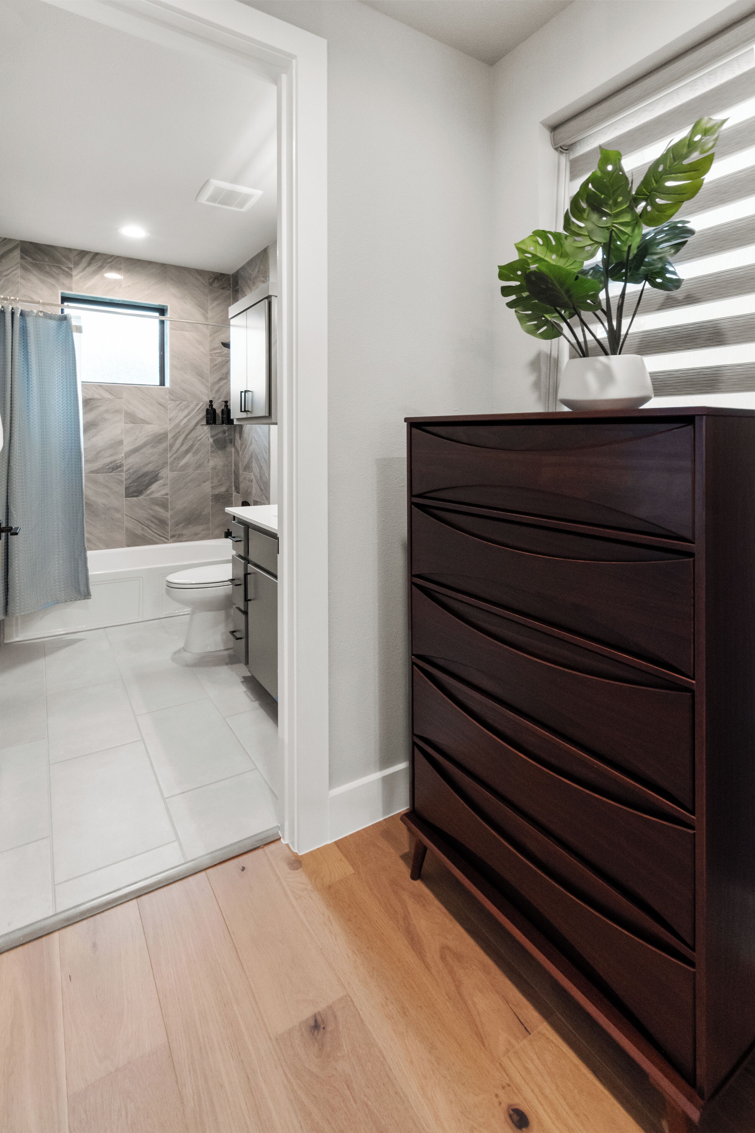 Un baño moderno con ducha, inodoro y lavamanos. Se aprecia una cómoda de madera oscura con cajones y una planta en su parte superior.