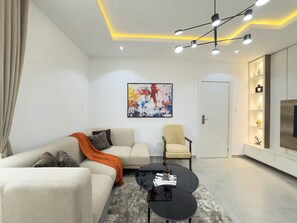 A modern living room with a sofa, armchair, and coffee table, a painting on the wall, and a door with a built-in TV. - Classic 1-Bedroom in Victoria Island with Pool, Fast Wi-Fi & 24/7 Power (Lagos)