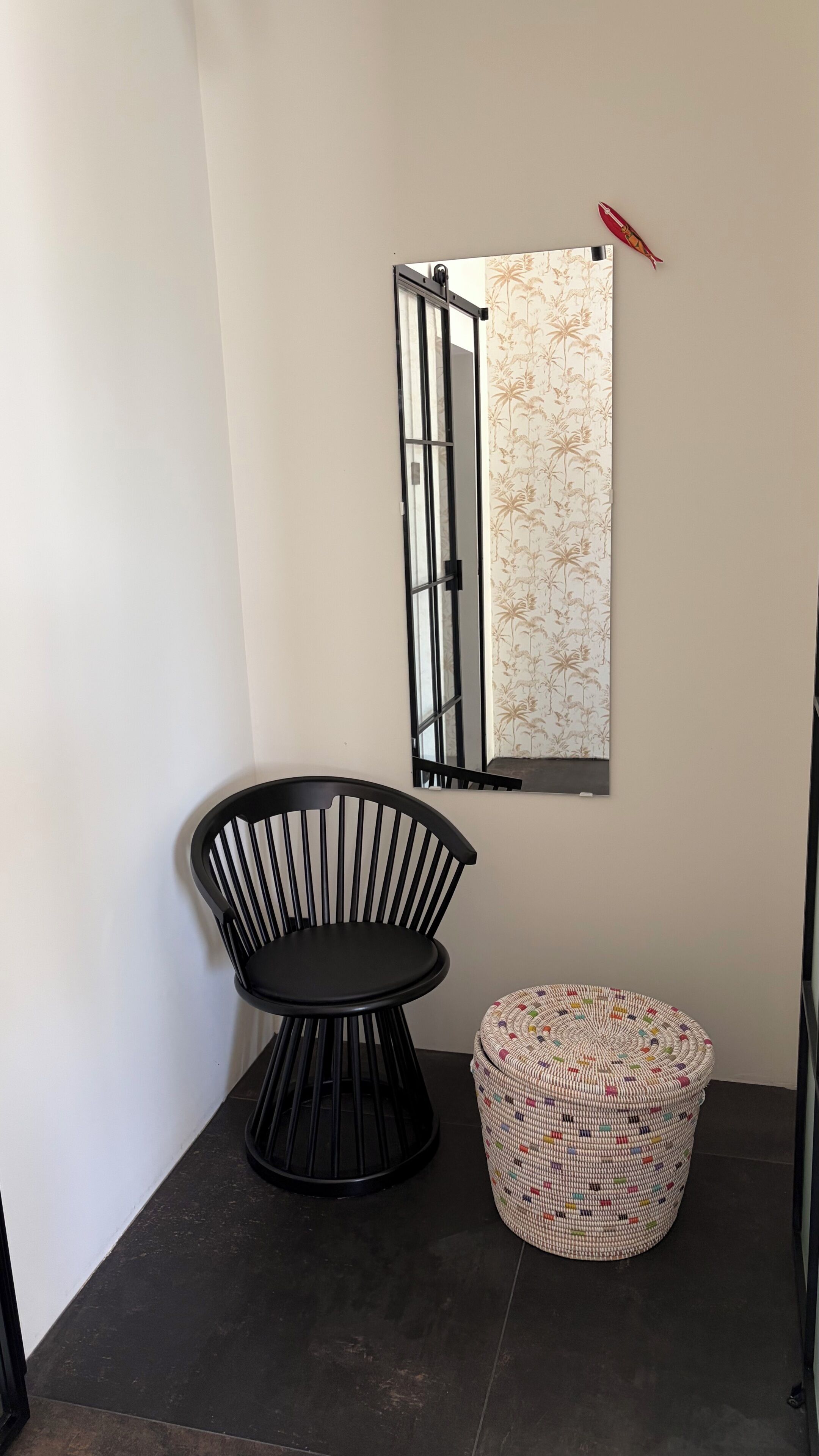 A corner room with a black chair, a woven ottoman, and a wall-mounted mirror with a floral pattern.