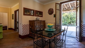 A dining area with a glass table, metal chairs, a bowl of fruit, and a view of the garden through an open door. - Villa Harmonia - Luxury Rooms - Salvia Suite - AC - By Guestaly (Dormelletto)