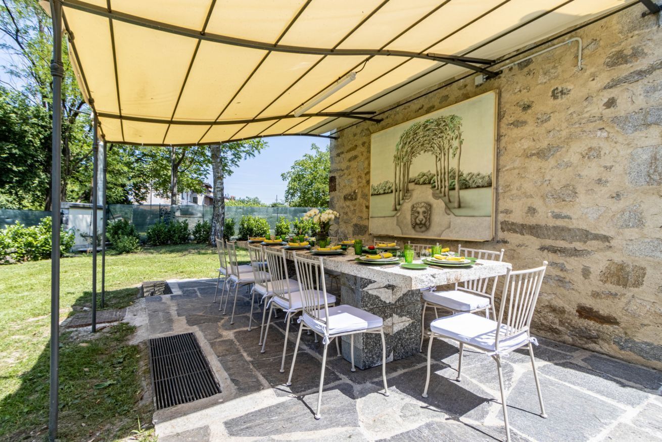 An outdoor dining area with a stone table and white chairs, a yellow canopy, and a stone wall with a painting.