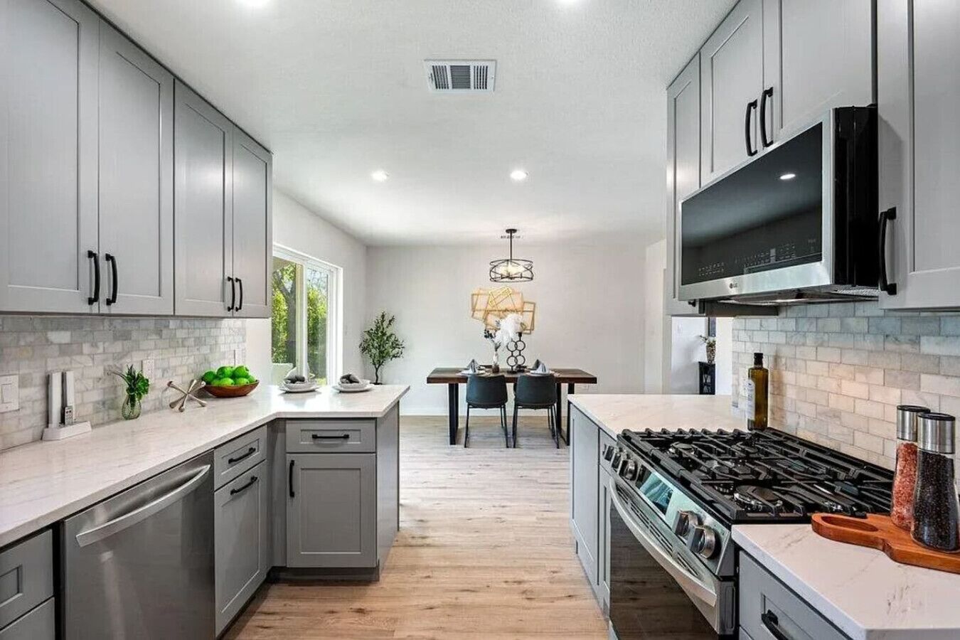 A modern kitchen with grey cabinets, a central island, a dining area with a chandelier, and a large TV above the stove.
