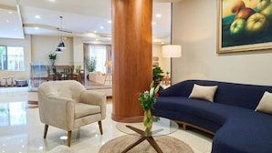 A modern hotel lobby with a round glass coffee table, a beige armchair, a blue sofa, and a large wooden pillar. - Suites Las Palmas Hotel & Apartments (San Salvador)