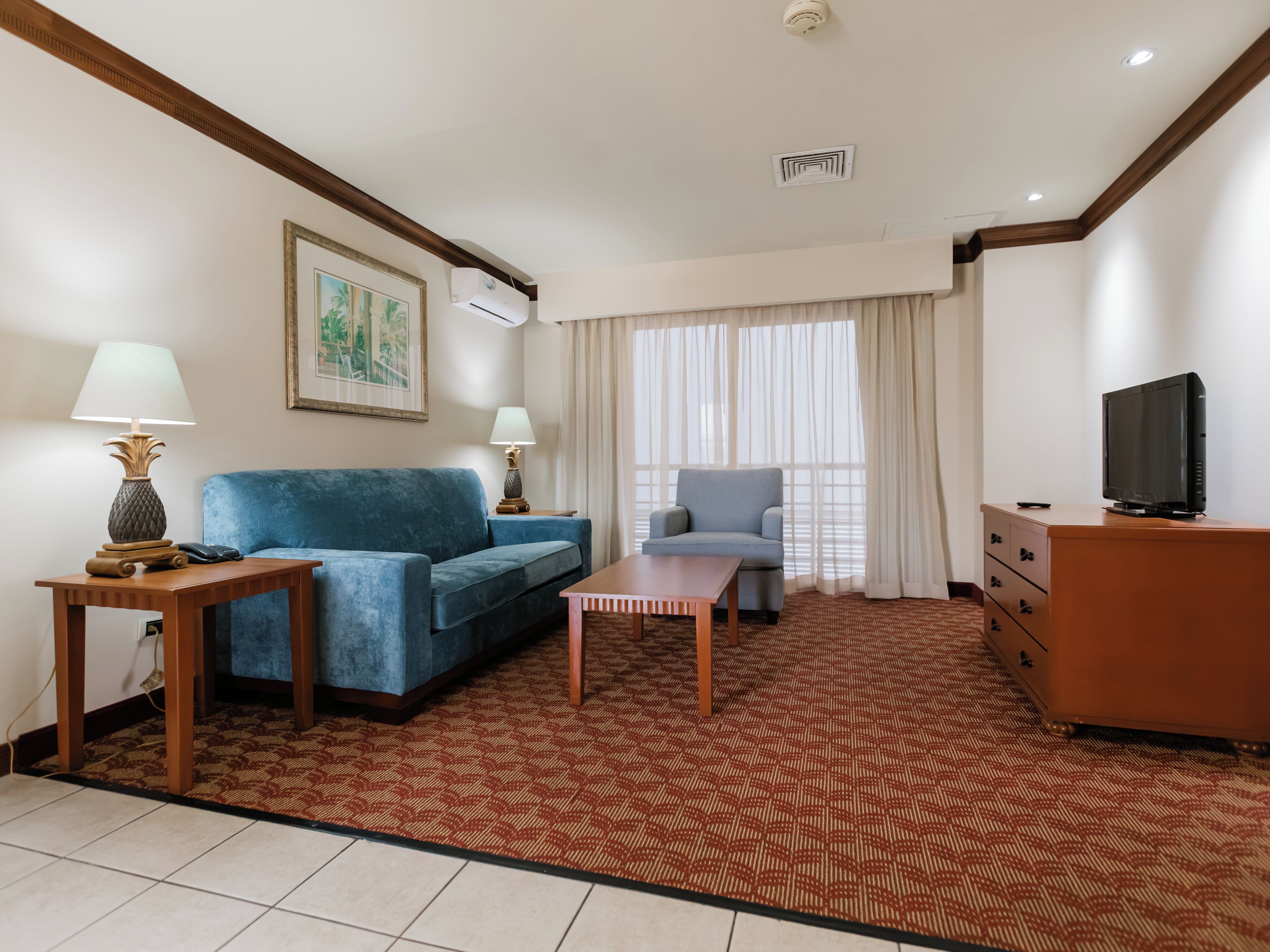 Quarto de hotel com um sofá azul, uma poltrona menor, uma mesa de madeira, uma televisão em uma cômoda e um tapete estampado.