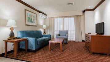 Quarto de hotel com um sofá azul, uma poltrona menor, uma mesa de madeira, uma televisão em uma cômoda e um tapete estampado.
