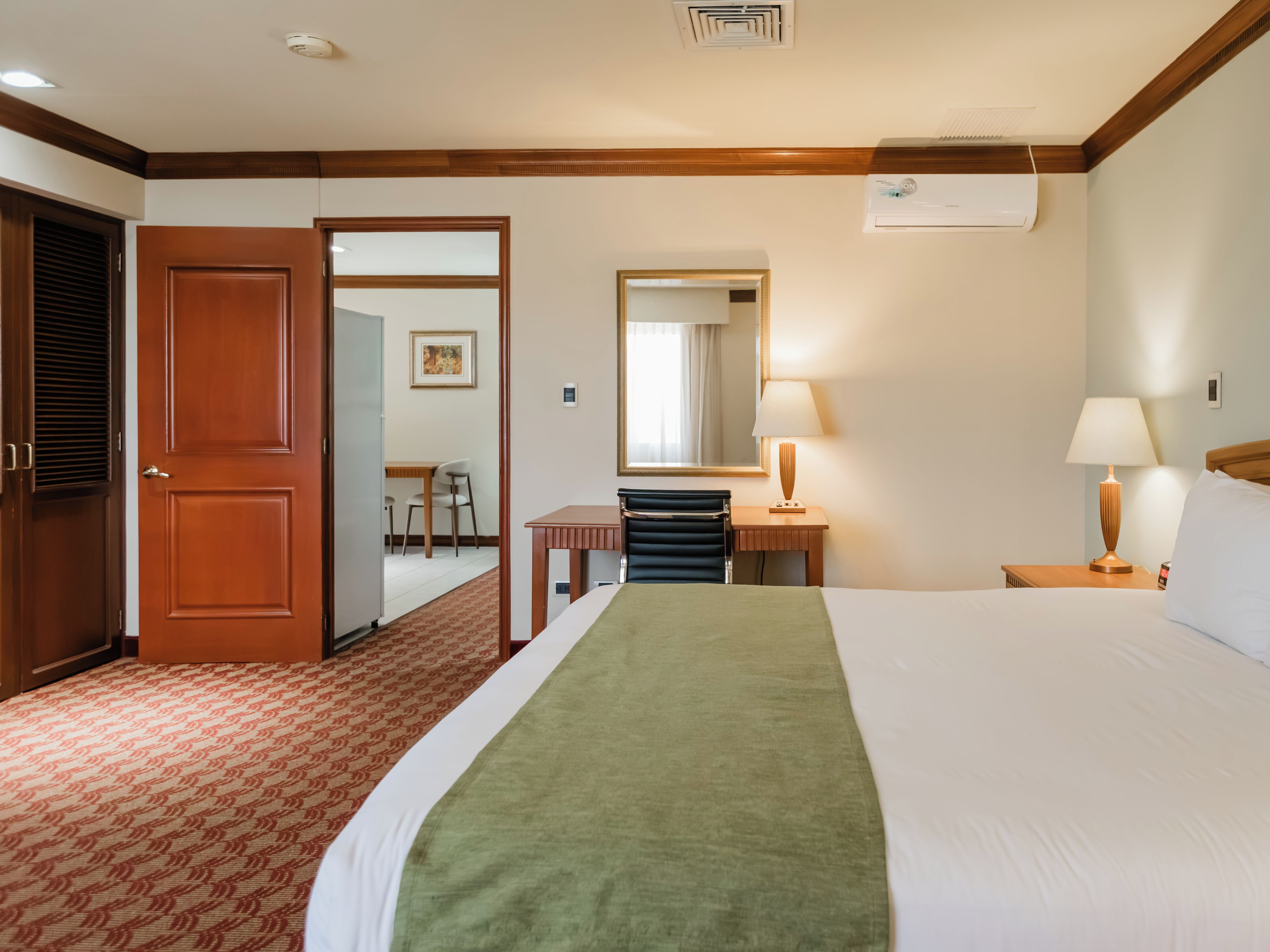Quarto de hotel com cama, mesas de cabeceira, espelho, porta e vista para o quarto vizinho.