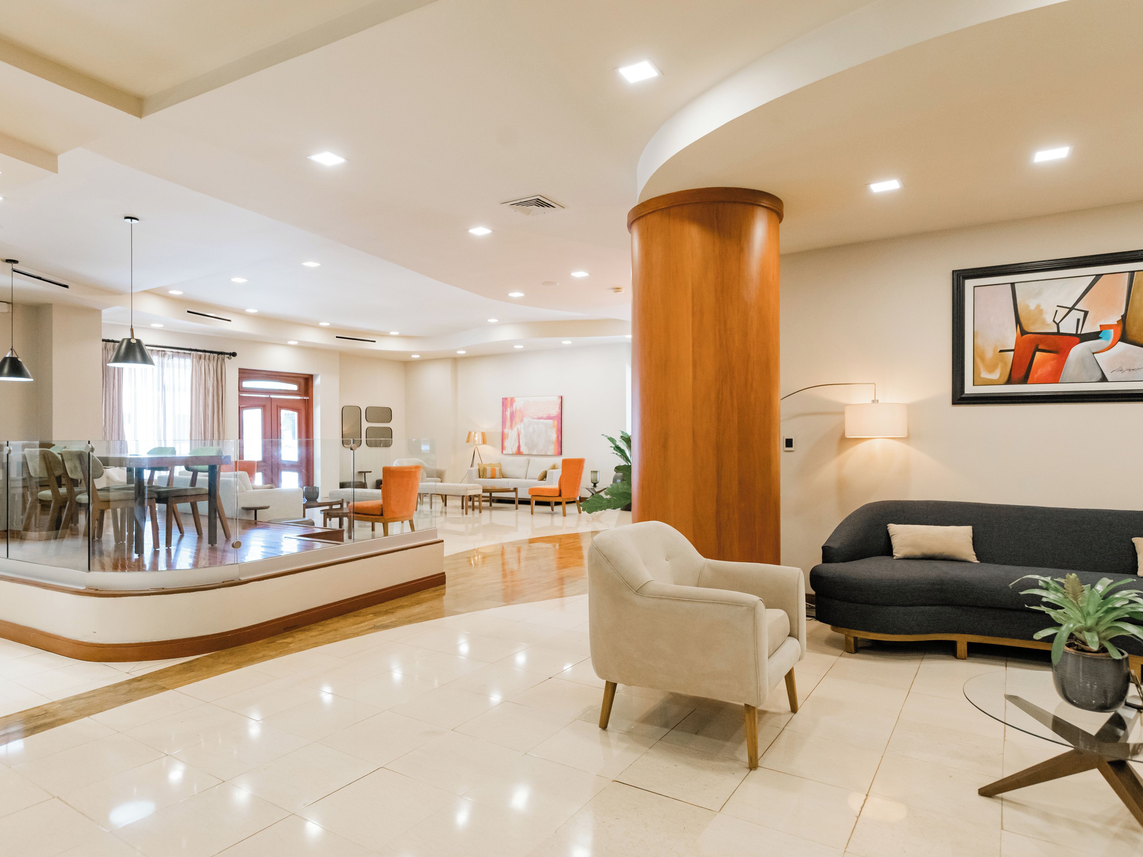 Un hall d’hôtel moderne avec un comptoir d’accueil incurvé, un espace salon avec des canapés et des fauteuils, un coin repas et une grande colonne en bois.