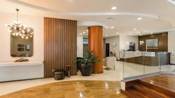 A modern hotel lobby with a reception desk, a large mirror, a wooden column, and a potted plant.