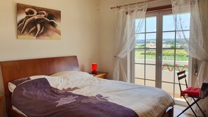 Ein Schlafzimmer mit einem Bett, einem Nachttisch, einer roten Lampe, einem Stuhl und Blick ins Freie durch ein Fenster mit Vorhängen.
