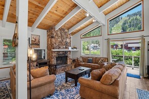 A spacious living room with a stone fireplace, wooden ceiling beams, and large windows offering a view of the outdoors. - Steps to Heavenly | Mountain Chalet w/ Hot Tub (South Lake Tahoe)