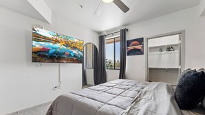 Ein Schlafzimmer mit einem Bett, einem an der Wand montierten Fernseher, einem Fenster mit Vorhängen und einer Tür mit Kleiderhaken.